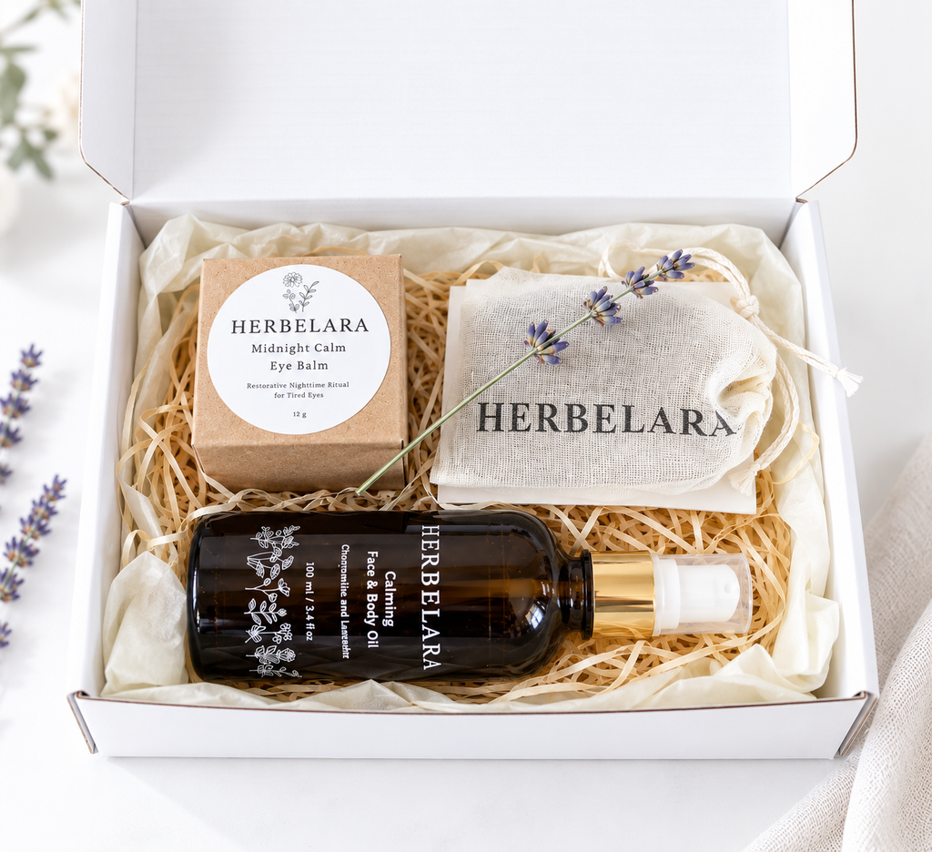 A curated eye care gift set from Herbelara arranged in a white box with shredded paper. The set includes a dark amber bottle of eye and face oil with a gold cap, a kraft paper box of herbal eye balm, a beige drawstring pouch, and a sprig of lavender.