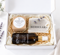A curated eye care gift set from Herbelara arranged in a white box with shredded paper. The set includes a dark amber bottle of eye and face oil with a gold cap, a kraft paper box of herbal eye balm, a beige drawstring pouch, and a sprig of lavender.