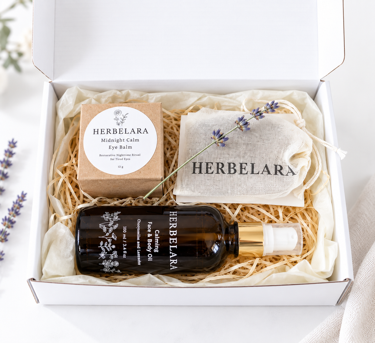A curated eye care gift set from Herbelara arranged in a white box with shredded paper. The set includes a dark amber bottle of eye and face oil with a gold cap, a kraft paper box of herbal eye balm, a beige drawstring pouch, and a sprig of lavender.